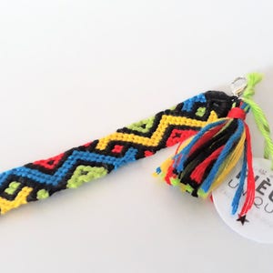May include: A colorful friendship bracelet with a black, yellow, red, and blue geometric pattern. The bracelet has a silver clasp and a multi-colored tassel. A white tag with black text reads "Pi&egrave;ce Unique".