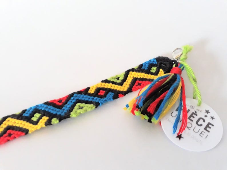May include: A colorful friendship bracelet with a black, yellow, red, and blue geometric pattern. The bracelet has a silver clasp and a multi-colored tassel. A white tag with black text reads "Pi&egrave;ce Unique".