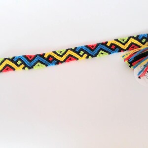 May include: A colorful friendship bracelet with a black, yellow, red, blue, and green geometric pattern. The bracelet has a tassel and a tag that says "PIECE UNIQUE" with stars.