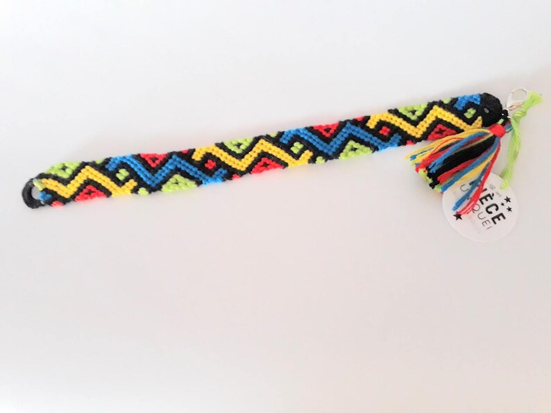 May include: A colorful friendship bracelet with a black, yellow, red, blue, and green geometric pattern. The bracelet has a tassel and a tag that says "PIECE UNIQUE" with stars.
