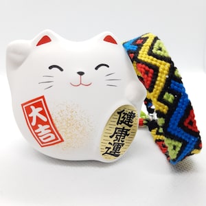 May include: White ceramic cat figurine with a smiling face and red and gold accents. The cat is holding a red sign with the text "大吉" and a gold sign with the text "錢庫豐盈". The cat is next to a colorful friendship bracelet with black, blue, red, yellow, and green stripes.