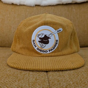 May include: Mustard yellow corduroy baseball cap featuring the San Diego Padres logo. The logo is a circular patch with a brown cartoon character swinging a baseball bat. The text "SAN DIEGO PADRES" is in a circular pattern around the character.