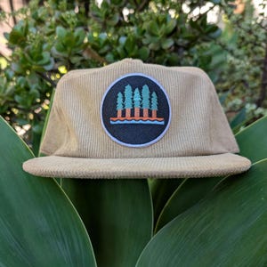 May include: Tan corduroy cap with a flat brim. A circular patch features a forest scene with green and orange trees, blue water, and a black background. The cap is set against a backdrop of green foliage.