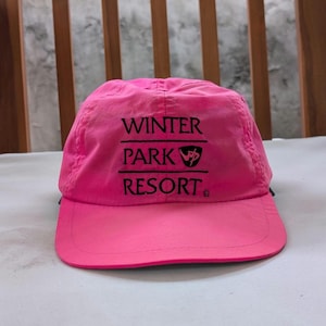 May include: A bright pink baseball cap with a curved brim. The cap features the text "WINTER PARK RESORT" in black, with a stylized logo. The cap is made of a lightweight material, suitable for outdoor activities.
