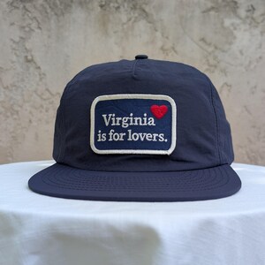 May include: A navy blue baseball cap featuring a rectangular patch on the front. The patch is navy blue with a white border and displays the white text Virginia is for lovers. with a small red embroidered heart above the word Virginia.