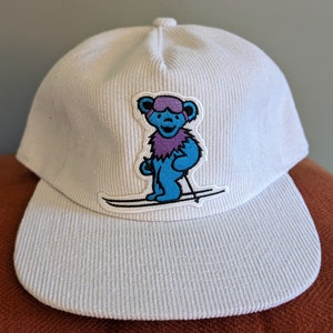 May include: White corduroy baseball cap featuring a colorful embroidered patch of a blue bear wearing goggles and a purple scarf, skiing on a pair of skis. The cap has a curved brim and a button on top.