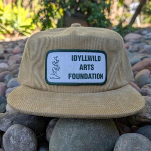 May include: A tan corduroy cap with a flat brim. The cap features a rectangular patch with the text "IDYLLWILD ARTS FOUNDATION" in white letters on a green background. A white graphic is on the left side of the patch.