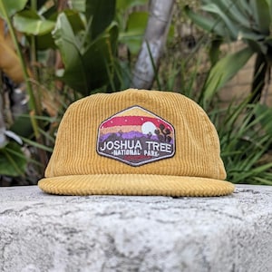 May include: A mustard yellow corduroy baseball cap featuring a hexagonal embroidered patch on the front. The patch depicts a desert landscape at sunset with a white sun, purple mountains, and the text Joshua Tree National Park.