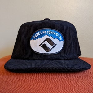 May include: Navy blue corduroy baseball cap with a flat brim. The cap features an embroidered patch with the text "THERE'S NO COMPARISON" above a mountain and logo design. The cap has a button on top.