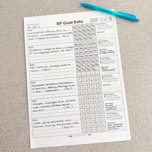 May include: A white paper IEP Goal Data tracking sheet with black printed text and handwritten entries, featuring a grid for progress monitoring. A bright teal ballpoint pen with the text The Intentional IEP rests on top.