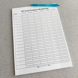 May include: A white paper sheet titled IEP Goal Progress Monitoring with a grid for tracking student data across six trials, featuring a column for student names and a teal ballpoint pen resting horizontally across the top edge of the page.