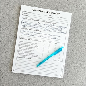 May include: A white Classroom Observation form with black text and lines for student, teacher, and class details. It includes sections for environment, accommodations, and a performance assessment grid, with a bright blue pen resting on top.