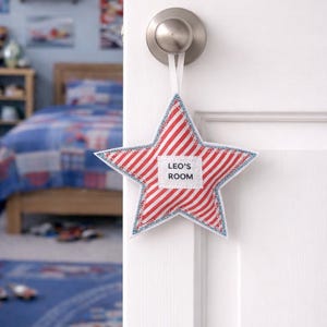 May include: A star-shaped door hanger with red and white stripes and a blue stitched border. The door hanger has a label that reads "LEO'S ROOM". It is hanging from a silver door knob on a white door.