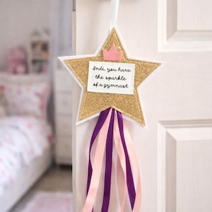 May include: A gold glitter star-shaped door hanger with a pink crown and ribbons. The text on the star reads, "Indi, you have the sparkle of a gymnast." The ribbons are pink and purple.
