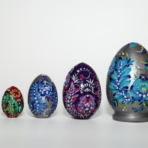May include: A collection of six decorative, hand-painted wooden Easter eggs. The eggs vary in size and feature floral designs in a range of colors, including blue, red, purple, and silver. The largest egg is approximately 6 inches tall.