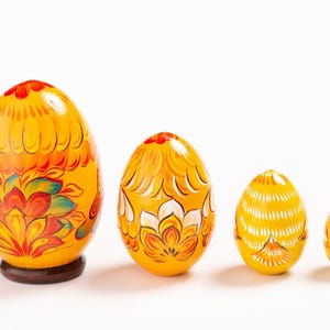 K&ouml;nnte beinhalten: Ein Set aus vier gelben, ineinander stapelbaren Holzeiern, die in einer Reihe von gro&szlig; nach klein angeordnet sind. Jedes Ei ist mit handgemalten Blumenmustern in Orange, Rot und Wei&szlig; verziert, wobei das gr&ouml;&szlig;te auf einem braunen Holzsockel steht.