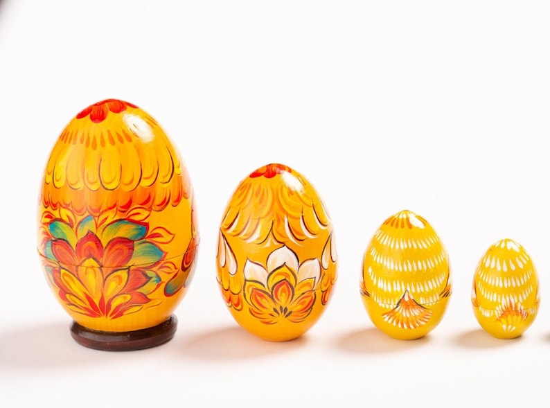 K&ouml;nnte beinhalten: Ein Set aus vier gelben, ineinander stapelbaren Holzeiern, die in einer Reihe von gro&szlig; nach klein angeordnet sind. Jedes Ei ist mit handgemalten Blumenmustern in Orange, Rot und Wei&szlig; verziert, wobei das gr&ouml;&szlig;te auf einem braunen Holzsockel steht.