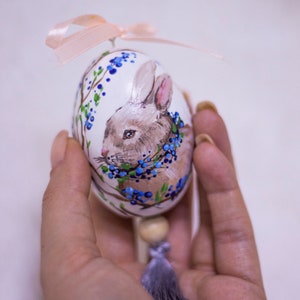 Puede incluir: Un huevo de Pascua pintado a mano con un conejo marrón y una guirnalda de bayas azules. El huevo blanco tiene detalles florales azules y verdes y una cinta rosa. Se adjuntan una cuenta de madera y una borla gris.