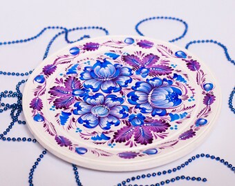 May include: A white, round decorative plate with a hand-painted floral design. The flowers are blue and purple, with green leaves. The plate is surrounded by a blue beaded garland.