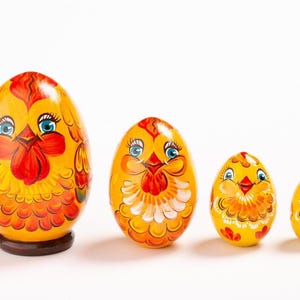 K&ouml;nnte beinhalten: Ein Set aus vier ineinander stapelbaren, eif&ouml;rmigen Holzfiguren in leuchtendem Gelb, Orange und Rot. Jede Figur zeigt ein handbemaltes H&uuml;hnergesicht mit blauen Augen, rotem Kamm und orangefarbenen Federn. Die gr&ouml;&szlig;te Figur steht auf einem dunkelbraunen Sockel.