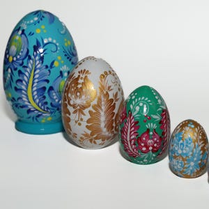 May include: A set of four nesting eggs arranged in a row, decreasing in size. The largest is bright blue with blue and yellow floral patterns. The second is white with gold leaf-like designs. The third is green with red and white floral motifs. The smallest is gold with blue flowers.