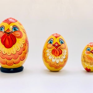 K&ouml;nnte beinhalten: Drei eif&ouml;rmige h&ouml;lzerne Matroschka-Puppen, bemalt als gelbe H&uuml;hner mit orangefarbenen und roten Federn, blauen Augen und orangefarbenen Schn&auml;beln. Die gr&ouml;&szlig;te Puppe steht auf einem schwarzen runden Sockel, die zwei kleineren haben abgerundete B&ouml;den.