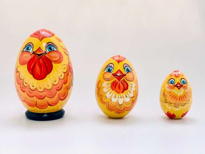 K&ouml;nnte beinhalten: Drei eif&ouml;rmige h&ouml;lzerne Matroschka-Puppen, bemalt als gelbe H&uuml;hner mit orangefarbenen und roten Federn, blauen Augen und orangefarbenen Schn&auml;beln. Die gr&ouml;&szlig;te Puppe steht auf einem schwarzen runden Sockel, die zwei kleineren haben abgerundete B&ouml;den.