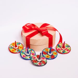 Puede incluir: Cinco peonzas de madera coloridas con dise&ntilde;os florales, dispuestas alrededor de una caja de regalo envuelta con una cinta roja.