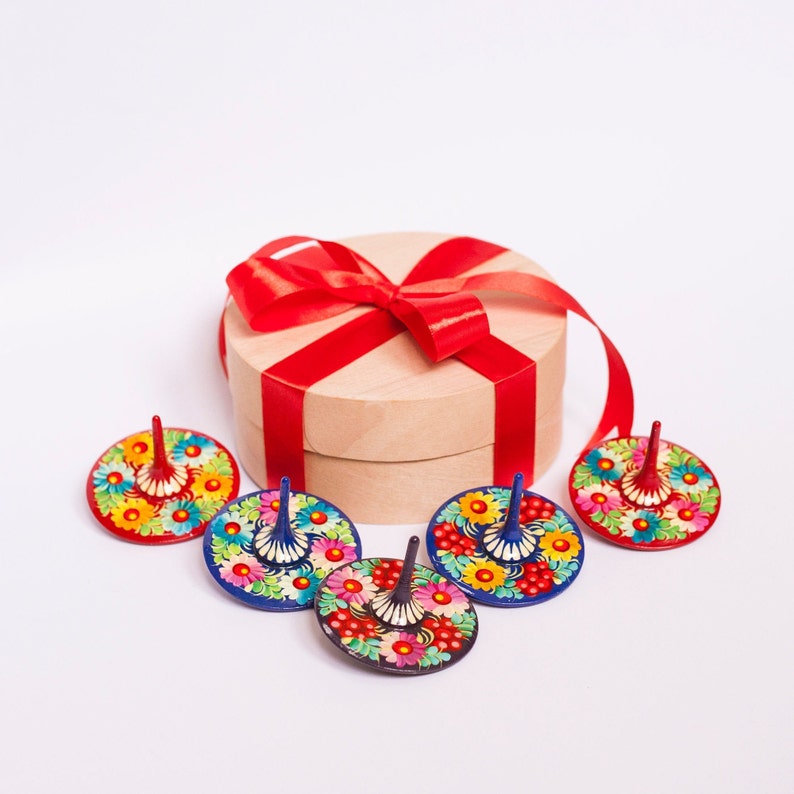 Puede incluir: Cinco peonzas de madera coloridas con dise&ntilde;os florales, dispuestas alrededor de una caja de regalo envuelta con una cinta roja.