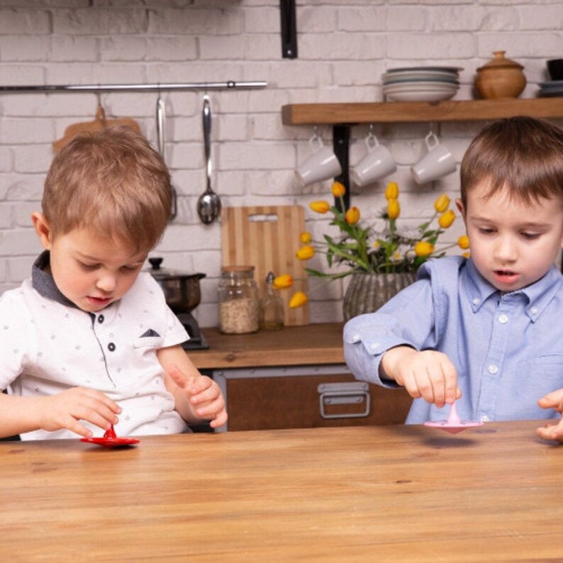 Puede incluir: Dos ni&ntilde;os est&aacute;n sentados en una mesa de madera, cada uno haciendo girar una peonza de colores. El ni&ntilde;o de la izquierda est&aacute; haciendo girar una peonza roja, mientras que el ni&ntilde;o de la derecha est&aacute; haciendo girar una peonza rosa.