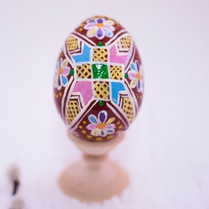Könnte beinhalten: Ein braunes Osterei mit einem geometrischen Muster, das in Rosa, Blau, Gelb und Grün bemalt ist. Das Ei ist mit kleinen weißen Blumen verziert und steht auf einem Holzständer.