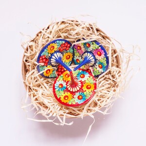 Puede incluir: Tres peonzas de madera coloridas con dise&ntilde;os florales azules, rojos, amarillos y blancos. Las peonzas est&aacute;n en una cesta llena de papel triturado.