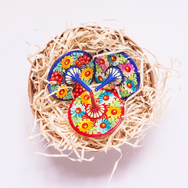 Puede incluir: Tres peonzas de madera coloridas con dise&ntilde;os florales azules, rojos, amarillos y blancos. Las peonzas est&aacute;n en una cesta llena de papel triturado.