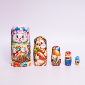 Könnte beinhalten: Ein Satz von fünf traditionellen russischen Nistpuppen, auch Matrjoschka-Puppen genannt, mit einem Hasenthema. Die größte Puppe ist mit einem weißen Hasen bemalt, der einen Korb mit bunten Eiern hält. Die anderen Puppen sind mit Hasen bemalt, die Karotten und Blumen halten.