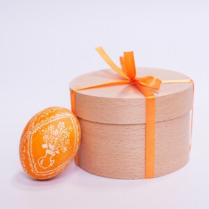 Könnte beinhalten: Eine runde Holzkiste mit hellbrauner Oberfläche ist mit einem leuchtend orangenen Band gebunden. Ein einzelnes orangefarbenes Osterei mit weißem Blumenmuster liegt neben der Kiste.