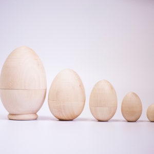May include: Five wooden eggs of varying sizes, arranged in a row from largest to smallest. The eggs are a light brown colour and have a smooth, polished finish.