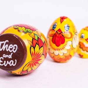 K&ouml;nnte beinhalten: Drei gelbe, eif&ouml;rmige Holzfiguren mit handgemalten Blumen- und H&uuml;hnermotiven. Das gr&ouml;&szlig;te Ei hat einen braunen Boden mit dem wei&szlig;en Schriftzug Theo and Eva. Das mittlere Ei zeigt ein H&uuml;hnergesicht und das kleinste Ei ist eine Miniaturversion.