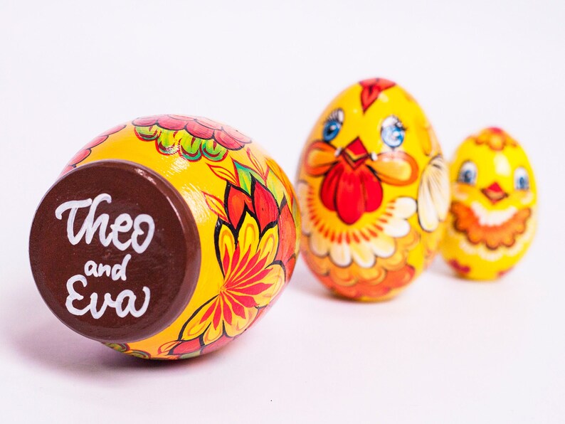 K&ouml;nnte beinhalten: Drei gelbe, eif&ouml;rmige Holzfiguren mit handgemalten Blumen- und H&uuml;hnermotiven. Das gr&ouml;&szlig;te Ei hat einen braunen Boden mit dem wei&szlig;en Schriftzug Theo and Eva. Das mittlere Ei zeigt ein H&uuml;hnergesicht und das kleinste Ei ist eine Miniaturversion.