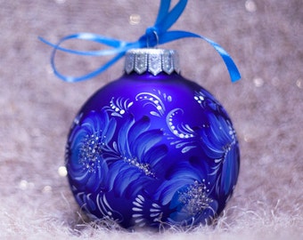 May include: A blue Christmas bauble with a white floral design painted on it. The bauble is hanging from a blue ribbon.