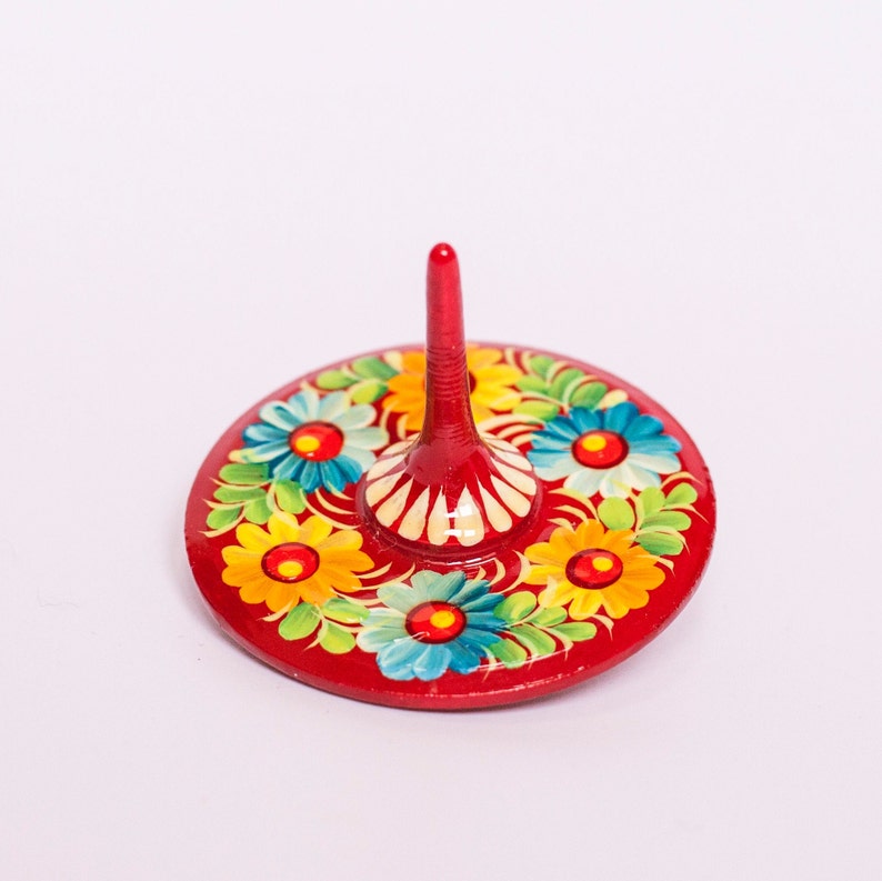 Puede incluir: Una peonza de madera roja con una parte superior puntiaguda y una base circular. La base est&aacute; decorada con flores coloridas en tonos de amarillo, azul, verde y rojo.