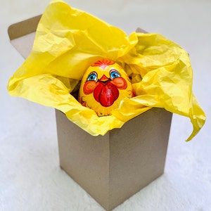 K&ouml;nnte beinhalten: Ein handbemaltes Holzei in Form eines Huhns mit gelbem K&ouml;rper, rotem Kamm und blauen Augen, eingebettet in eine braune Pappschachtel, die mit hellgelbem Seidenpapier ausgekleidet ist. Das Huhn zeigt detaillierte Pinselstriche und einen fr&ouml;hlichen Ausdruck.