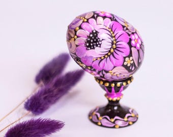 Op de afbeelding: Een handbeschilderd paasei met een bloemmotief in roze en gouden tinten op een zwarte achtergrond. Het ei wordt gepresenteerd op een zwarte standaard met gouden accenten.