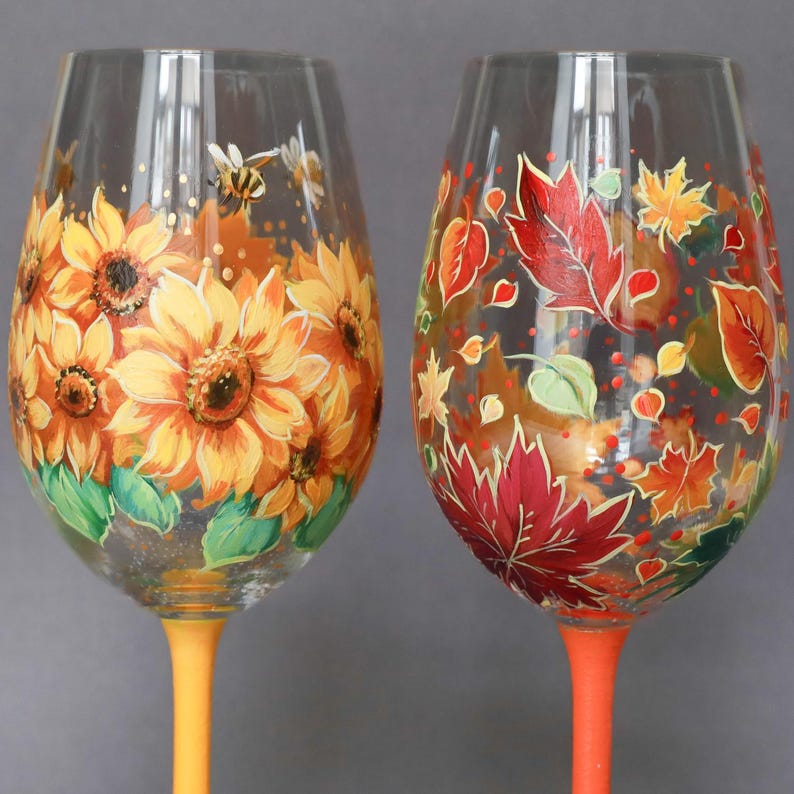 May include: Two hand-painted wine glasses. One features sunflowers and a bee, the other autumn leaves. The stems are painted yellow and orange, respectively. The glasses are clear with colorful floral designs.