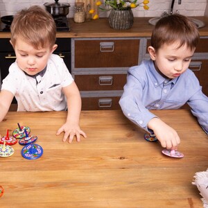 Puede incluir: Dos ni&ntilde;os juegan con peonzas coloridas en una mesa de madera. Las peonzas tienen dise&ntilde;os intrincados y est&aacute;n pintadas en tonos de azul, rojo, amarillo y verde.