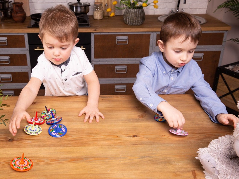 Puede incluir: Dos ni&ntilde;os juegan con peonzas coloridas en una mesa de madera. Las peonzas tienen dise&ntilde;os intrincados y est&aacute;n pintadas en tonos de azul, rojo, amarillo y verde.