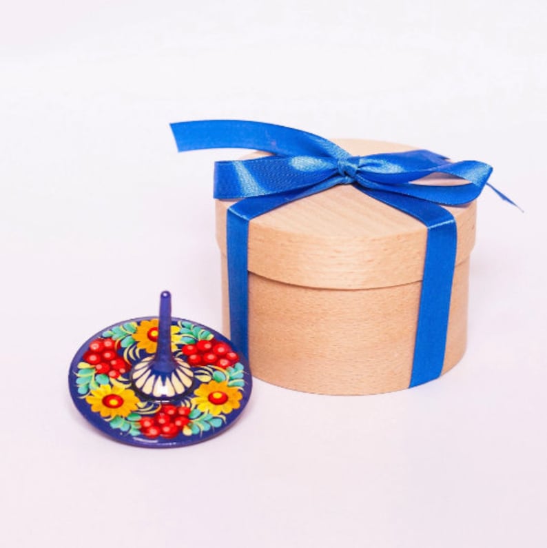 Puede incluir: Un peque&ntilde;o trompo de madera pintado de azul y rojo con un dise&ntilde;o floral. El trompo est&aacute; sentado junto a una caja de regalo redonda de madera con una cinta azul.
