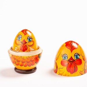 K&ouml;nnte beinhalten: Ein h&ouml;lzernes Nesting-Ei, bemalt als gelbes Huhn mit orangefarbenen Federn und blauen Augen. Das Ei ist in zwei Teile getrennt und enth&uuml;llt ein kleineres, passendes gelbes H&uuml;hnerei in der Basis, das auf einem dunkelbraunen, runden Sockel steht.