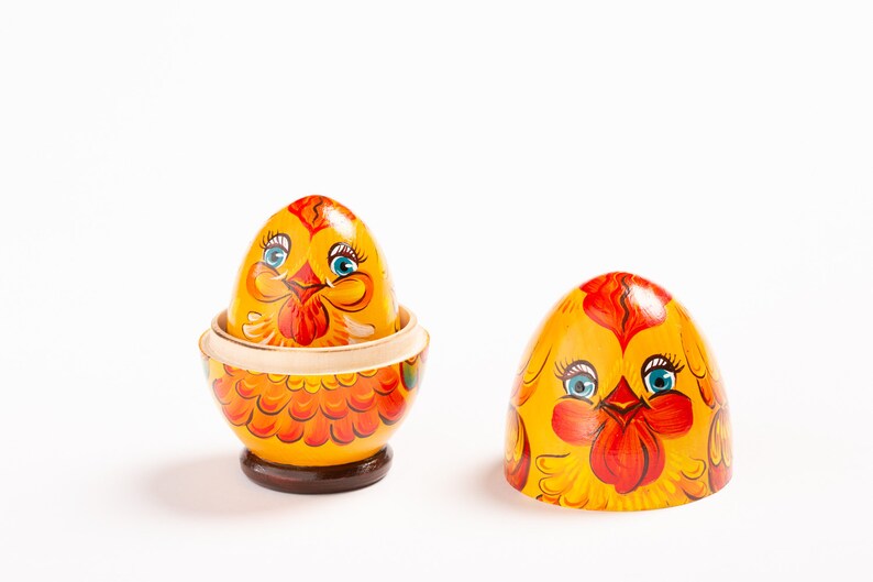 K&ouml;nnte beinhalten: Ein h&ouml;lzernes Nesting-Ei, bemalt als gelbes Huhn mit orangefarbenen Federn und blauen Augen. Das Ei ist in zwei Teile getrennt und enth&uuml;llt ein kleineres, passendes gelbes H&uuml;hnerei in der Basis, das auf einem dunkelbraunen, runden Sockel steht.