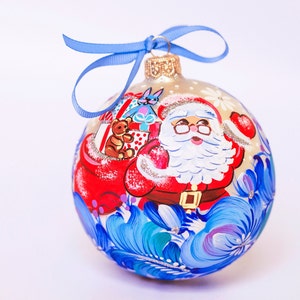 Könnte beinhalten: Ein handbemalter Weihnachtsbaumschmuck mit dem Weihnachtsmann, der einen Teddybär und Geschenke trägt. Der Schmuck ist mit blauen und weißen Blumendesigns und einem hellgoldenen Band verziert.