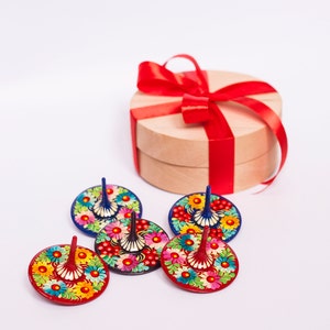 Puede incluir: Cinco peonzas de madera coloridas con dise&ntilde;os florales. Las peonzas est&aacute;n dispuestas en c&iacute;rculo alrededor de una caja de regalo redonda con una cinta roja.
